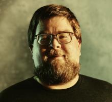 The head of rotund, bearded man with short, brown hair and glasses lit mostly from the right side.